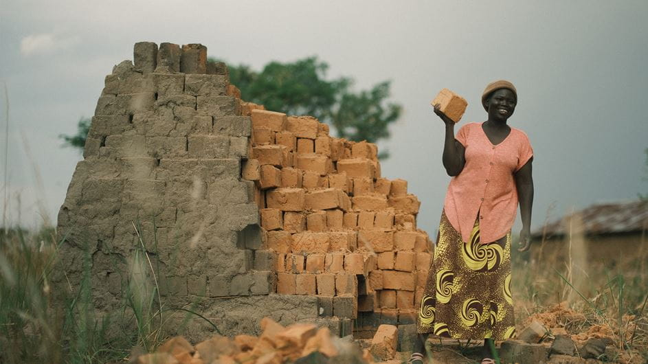 A woman is smiling as she stands beside a pile of bricks, holding a single brick.